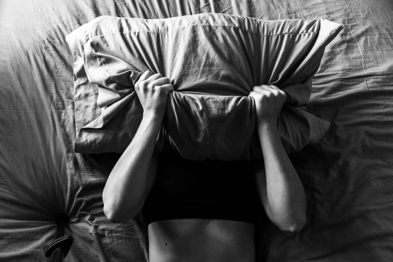 Black and white photograph of a person gripping a pillow that is over their face.